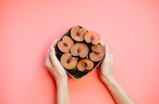 Top view of halved plums in a bowl held by hands on a vibrant pink background.