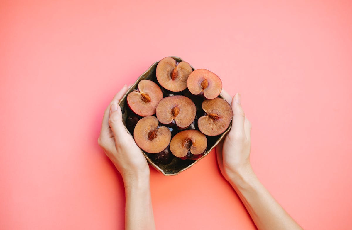 Crop faceless lady demonstrating bowl of ripe halved plums against pink background