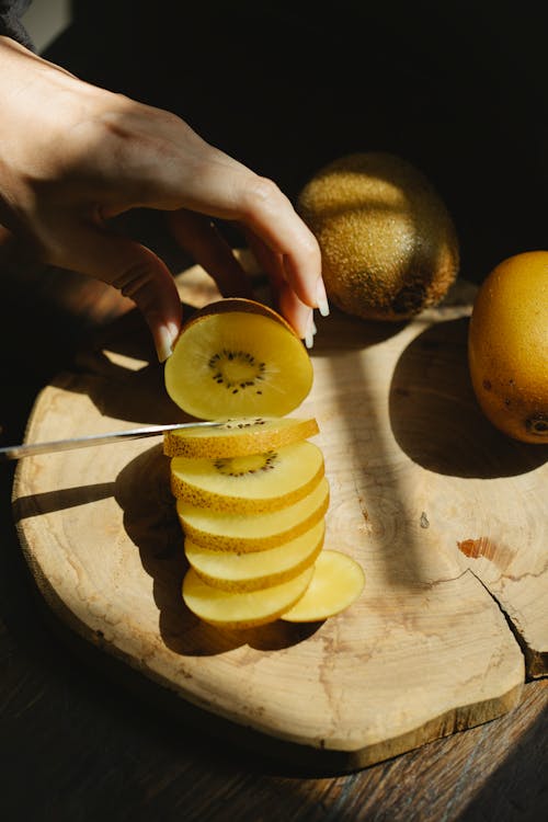 From above crop anonymous female cutting fresh ripe kiwi on wooden chopping board for healthy meal