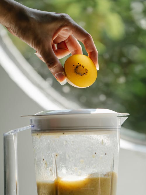 Crop anonymous female adding fresh fruit to blender with smoothie while cooking healthy food in kitchen