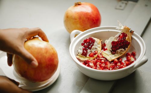 Free Unrecognizable person standing in kitchen at gray table with wholesome pomegranate and white bowl full of red seeds and peel Stock Photo