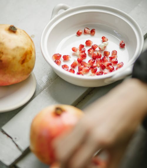 Unrecognizable blurred person putting removed seeds of pomegranate from flesh and peel into white bowl on gray table