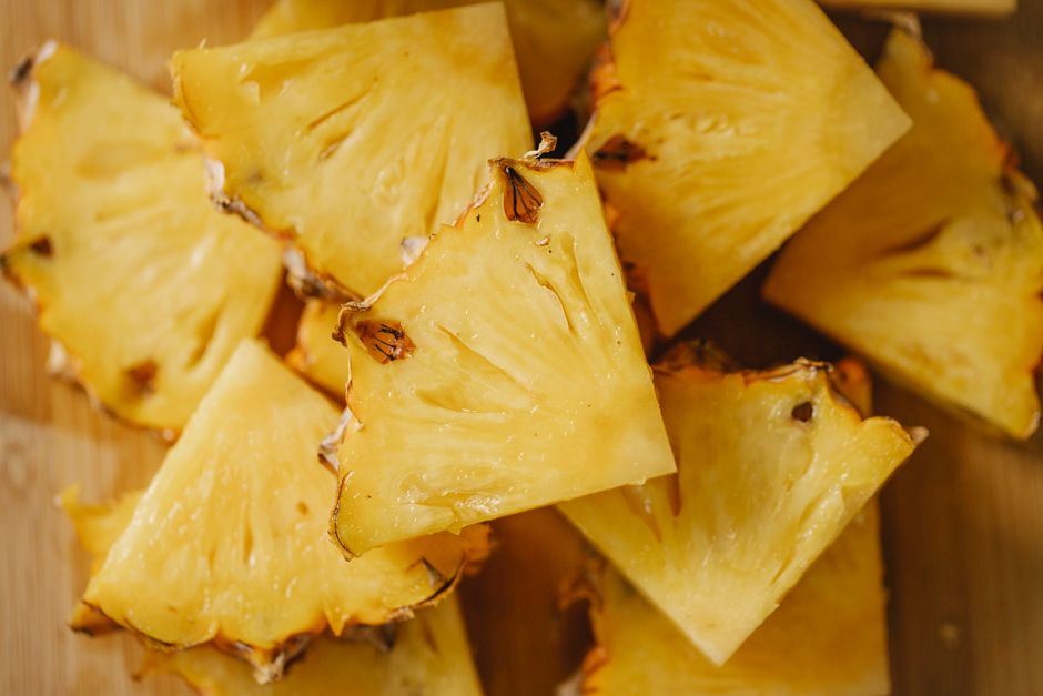 A whole pineapple with its crown and rind removed, sliced into rings on a wooden cutting board next to a pile of cored pineapple chunks.