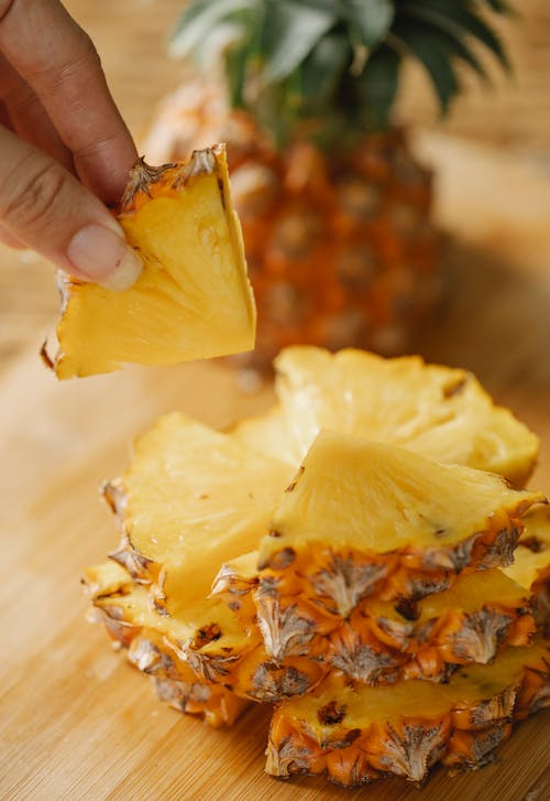 Unrecognizable female taking triangular segment of ripe pineapple placed on cutting board with half of fruit on blurred background in kitchen
