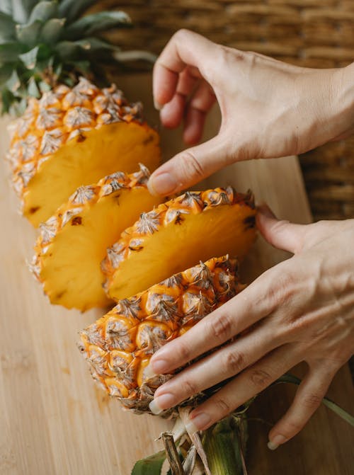 Free From above of anonymous female arranging round shaped exotic pineapple pieces on cutting board near half of fruit with green crown in kitchen Stock Photo