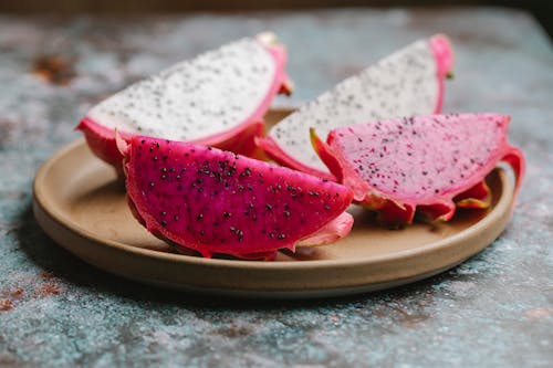 Ripe segments of dragon fruit with black seeds in colorful flesh served on round plate on gray table in kitchen on blurred background