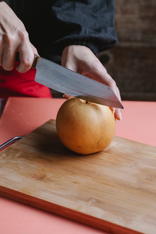 Unrecognizable person with sharp knife cutting ripe snow pear on cutting board while standing at table in kitchen during cooking