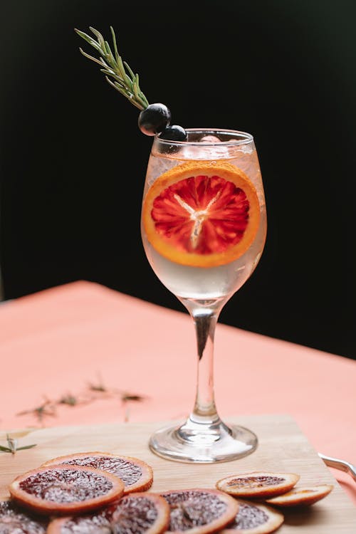 Wineglass with cold transparent drink decorated with olives on rosemary placed on cutting board with slices of blood orange on blurred black background