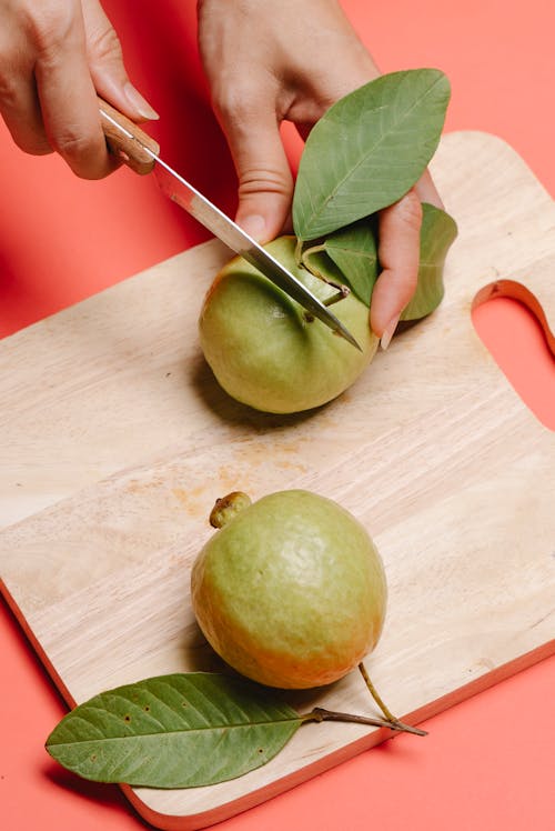 High angle of crop anonymous female with sharp knife cutting fresh ripe guava in half on wooden chopping board on pink background