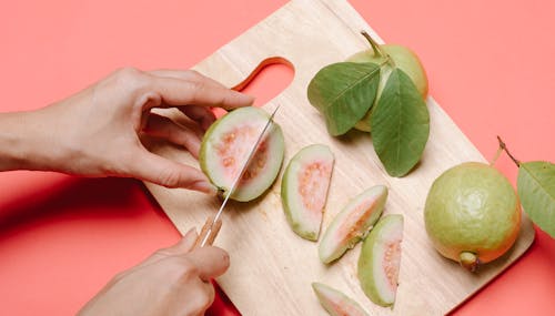 From above crop anonymous female with knife cutting fresh ripe fruits of guava on wooden chopping board on pink background