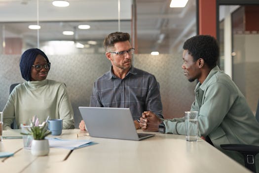 Three colleagues engaged in a discussion around a laptop, showcasing teamwork and diversity in a contemporary office setting.