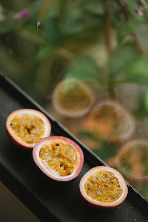From above several ripe juicy appetizing halves of passion fruits placed in row on black windowsill and green plants behind glass