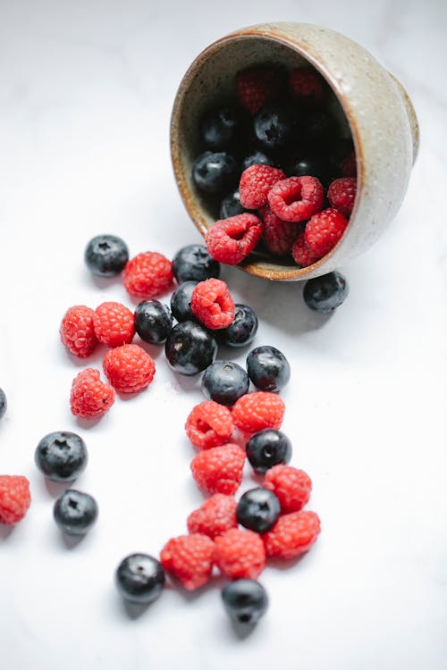 From above composition of ripe yummy raspberries and blueberries from bowl scattered on white table