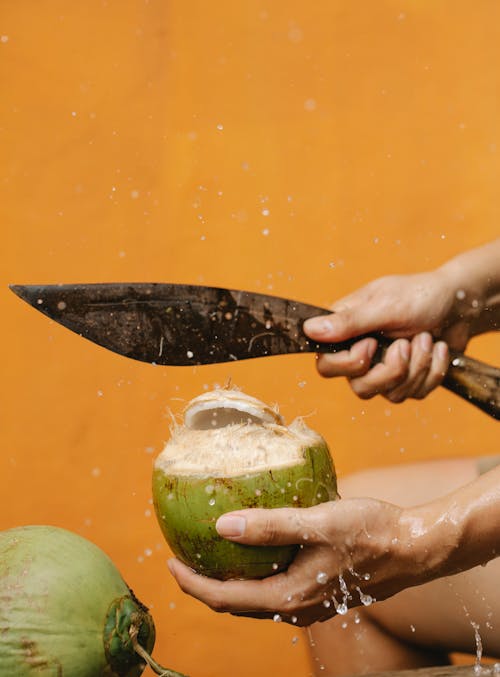 Side view of crop anonymous male cutting fresh coconut with sharp knife against orange background