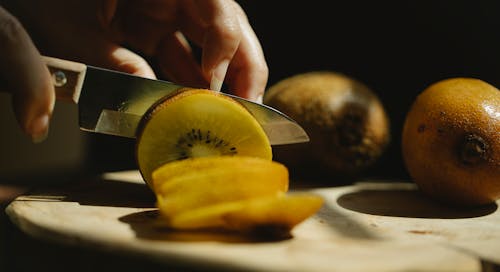 Closeup of crop unrecognizable female cutting fresh ripe kiwi with small sharp knife on wooden chopping board