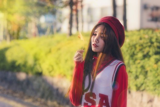 Young woman in a red sweater and beret holds a leaf outdoors, bathed in warm autumn light.