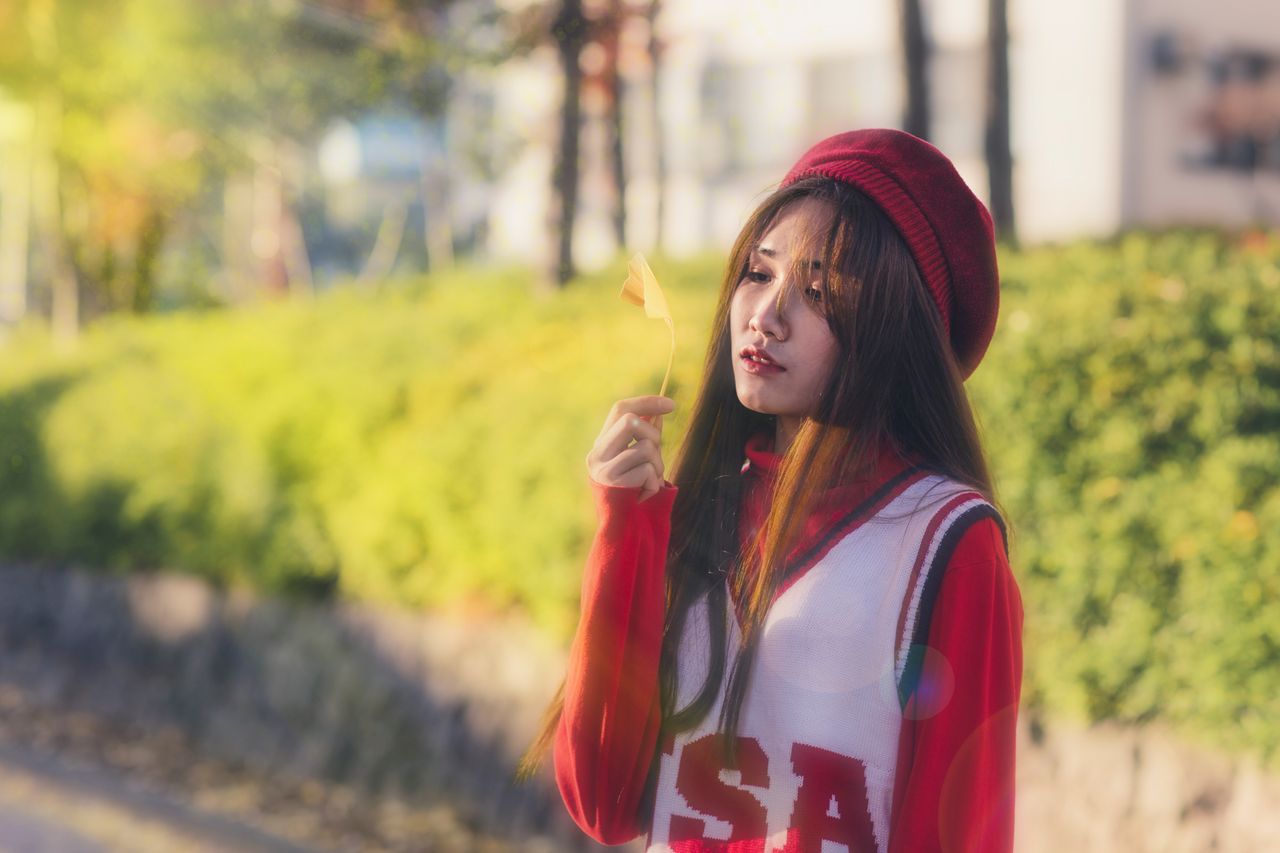 young woman in a red sweater and beret holds a leaf outdoors bathed in