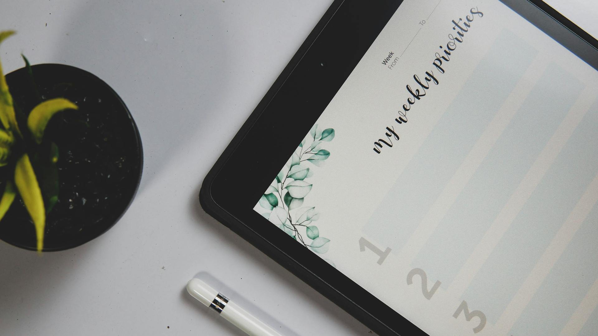 Close-up of a tablet displaying a weekly planner with a stylus and a potted plant on a desk.