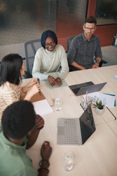 A diverse group of colleagues engaged in a meeting at a modern office, focusing and discussing with laptops.