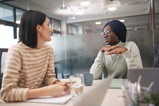 Two female colleagues engaged in a friendly discussion in a modern office setting.