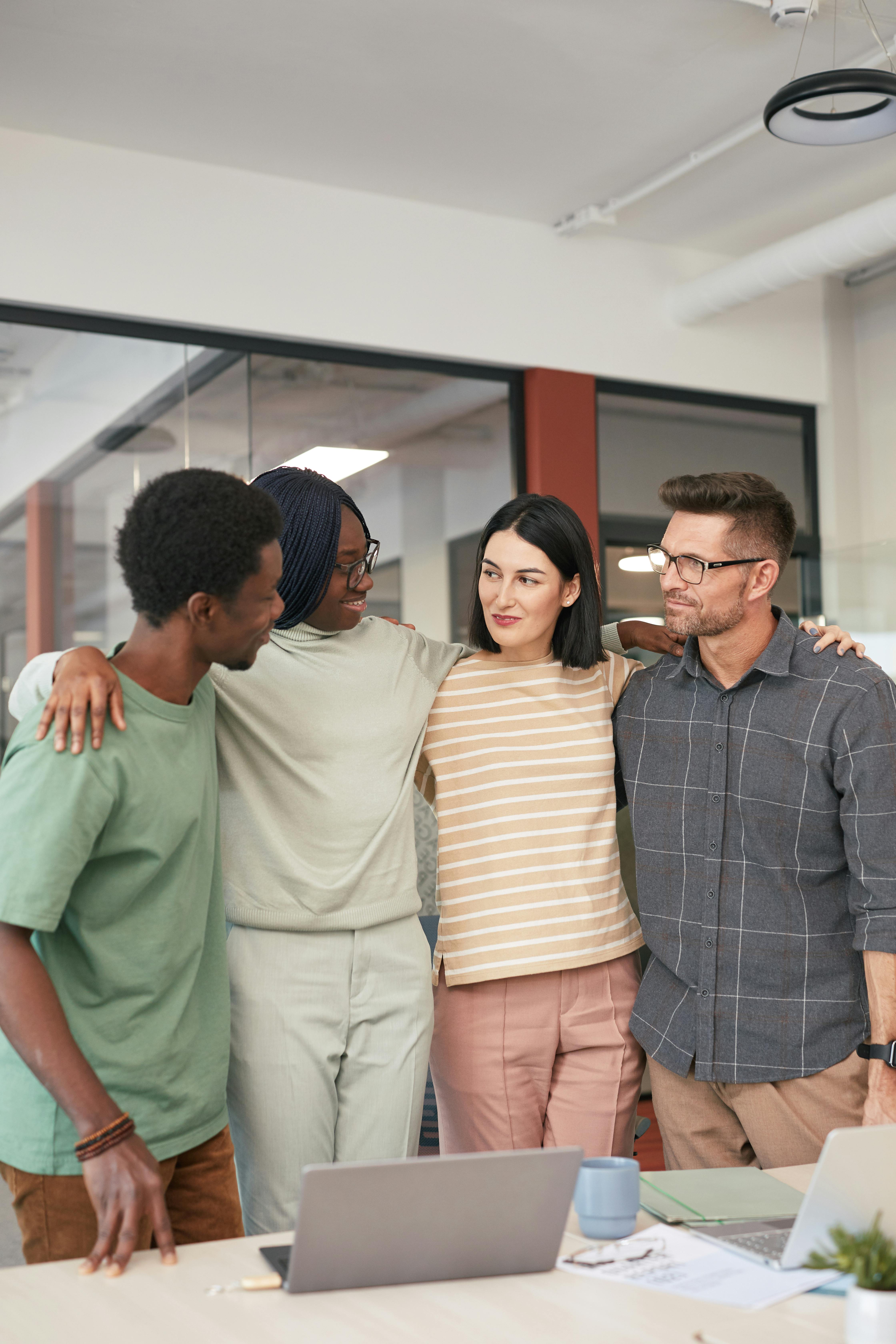A diverse group of colleagues standing with arms around each other at a modern office.