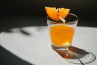 Glass of alcohol drink decorated with slice of orange on wooden stick placed on white table on blurred black background