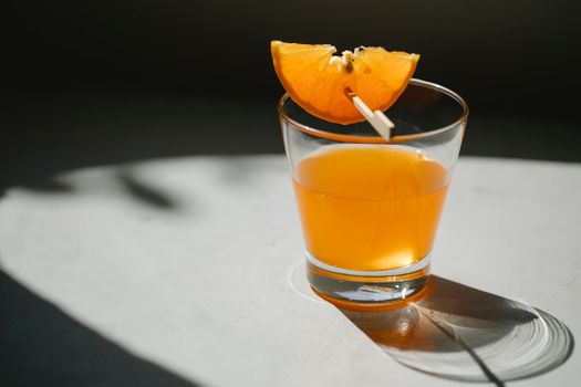 Glass of alcohol drink decorated with slice of orange on wooden stick placed on white table on blurred black background