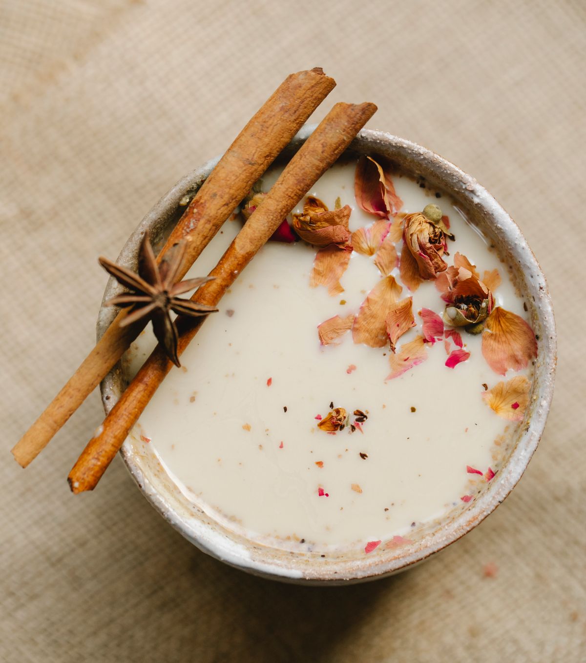 Overhead view of creamy spiced milk topped with dried rose petals, cinnamon sticks and star anise