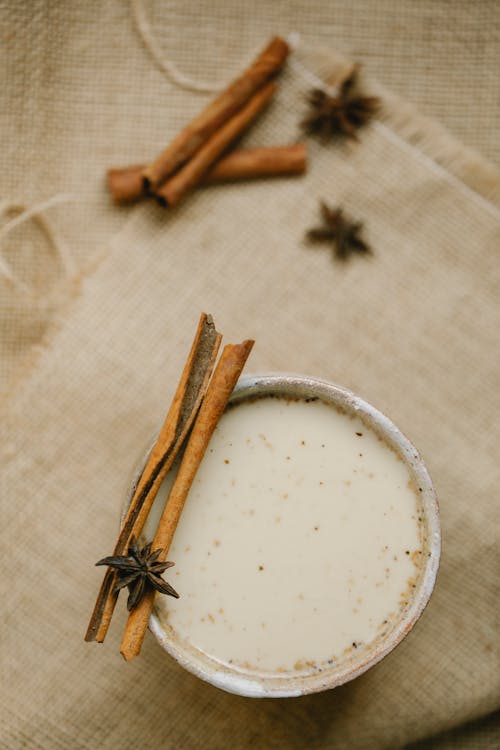 Top view mug of masala chai with milk decorated with cinnamon sticks and anise stars placed on fabric on blurred background