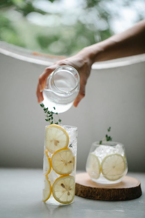 Unrecognizable person pouring water from jug into glass with lemon slices placed on table near drink with ice cubes in room with round window on blurred background