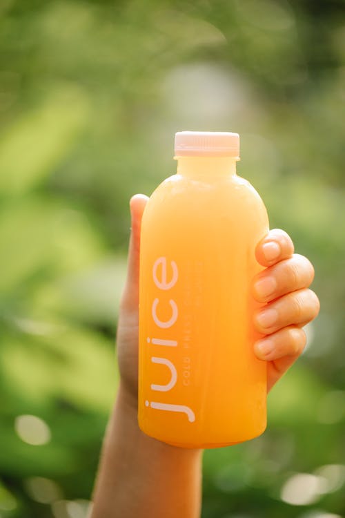 Unrecognizable person demonstrating bottle of orange beverage while standing in nature with green foliage on blurred background on sunny day