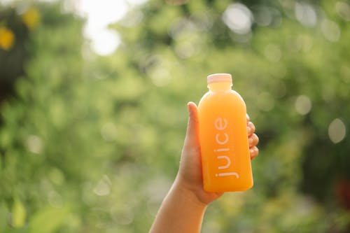 Unrecognizable person demonstrating bottle of orange juice while standing in nature with green trees and lush foliage on blurred background