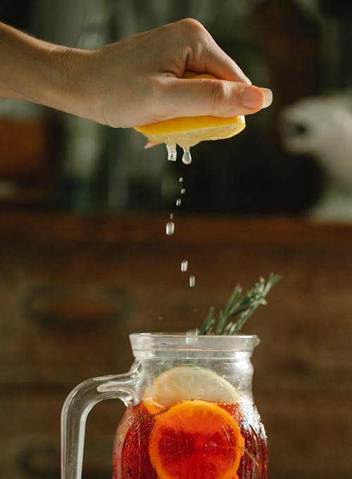 Unrecognizable female squeezing juice into glass jug with red cocktail and lemon slices decorated with rosemary in kitchen on blurred background