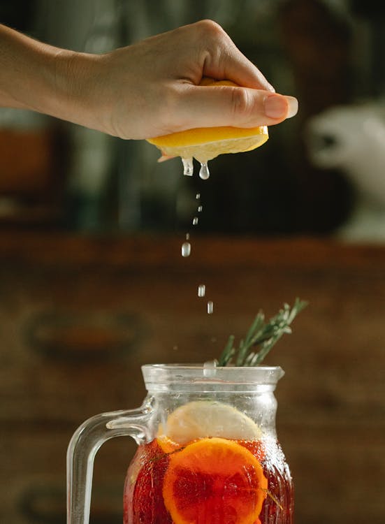 Free Unrecognizable female squeezing juice into glass jug with red cocktail and lemon slices decorated with rosemary in kitchen on blurred background Stock Photo