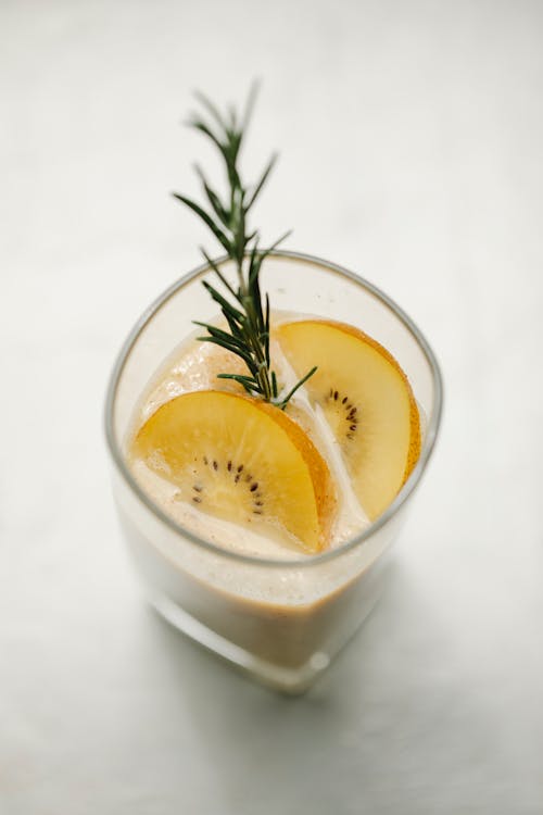 From above glass of fruit smoothie decorated with kiwi slices and rosemary sprig placed on white background in light kitchen