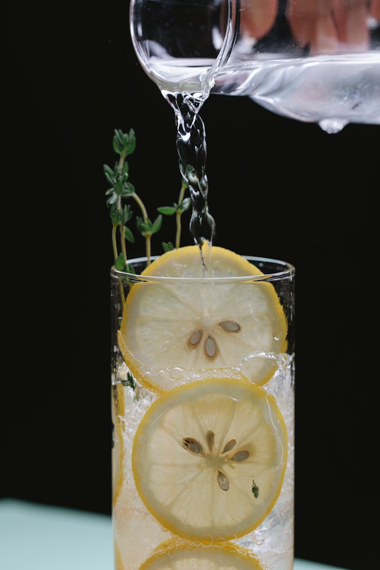 Person Pouring Liquid From Glass Jug Into Glass With Lemon