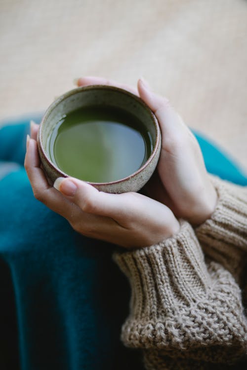 From above of crop anonymous female in knitwear sitting on floor with cup of matcha tea