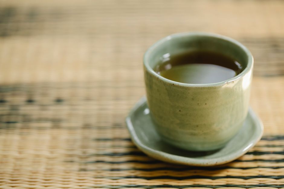 ウィッカーマットの上にある緑の陶器のカップに入った蒸気の立つお茶のミニマリストな構成は、静けさと伝統の本質を捉えています。