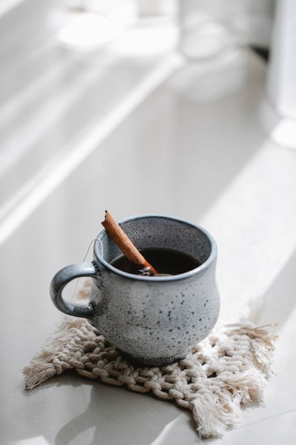 From above of ceramic cup with hot aromatic tea with cinnamon stick on knitted coaster on white table near window in sunlight