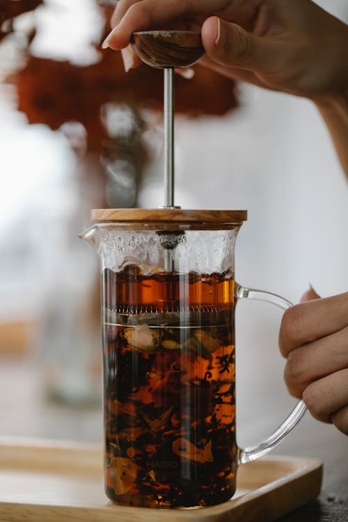 Free Crop anonymous female touching glass French press teapot with brewing aromatic black tea placed on tray Stock Photo
