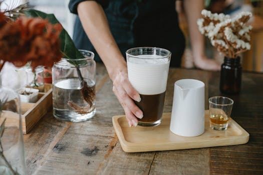 Crop anonymous female barista serving fresh brewed iced chocolate coffee with thick layer of fluffy froth on wooden tray with shot of cognac