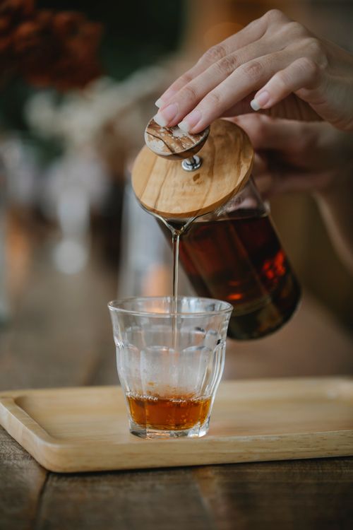 Free Crop anonymous female pouring hot black tea from French press teapot into glass placed on wooden tray Stock Photo