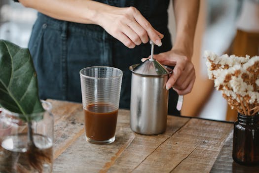Crop anonymous female barista making froth in manual steel milk frother while preparing iced chocolate coffee in light kitchen