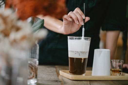 Close-up of a barista stirring a creamy coffee beverage with a frothy top in a café setting.