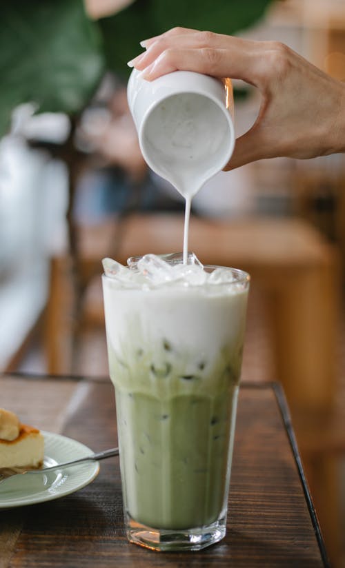Crop unrecognizable female barista adding fresh milk from jigger into glass of yummy iced matcha latte