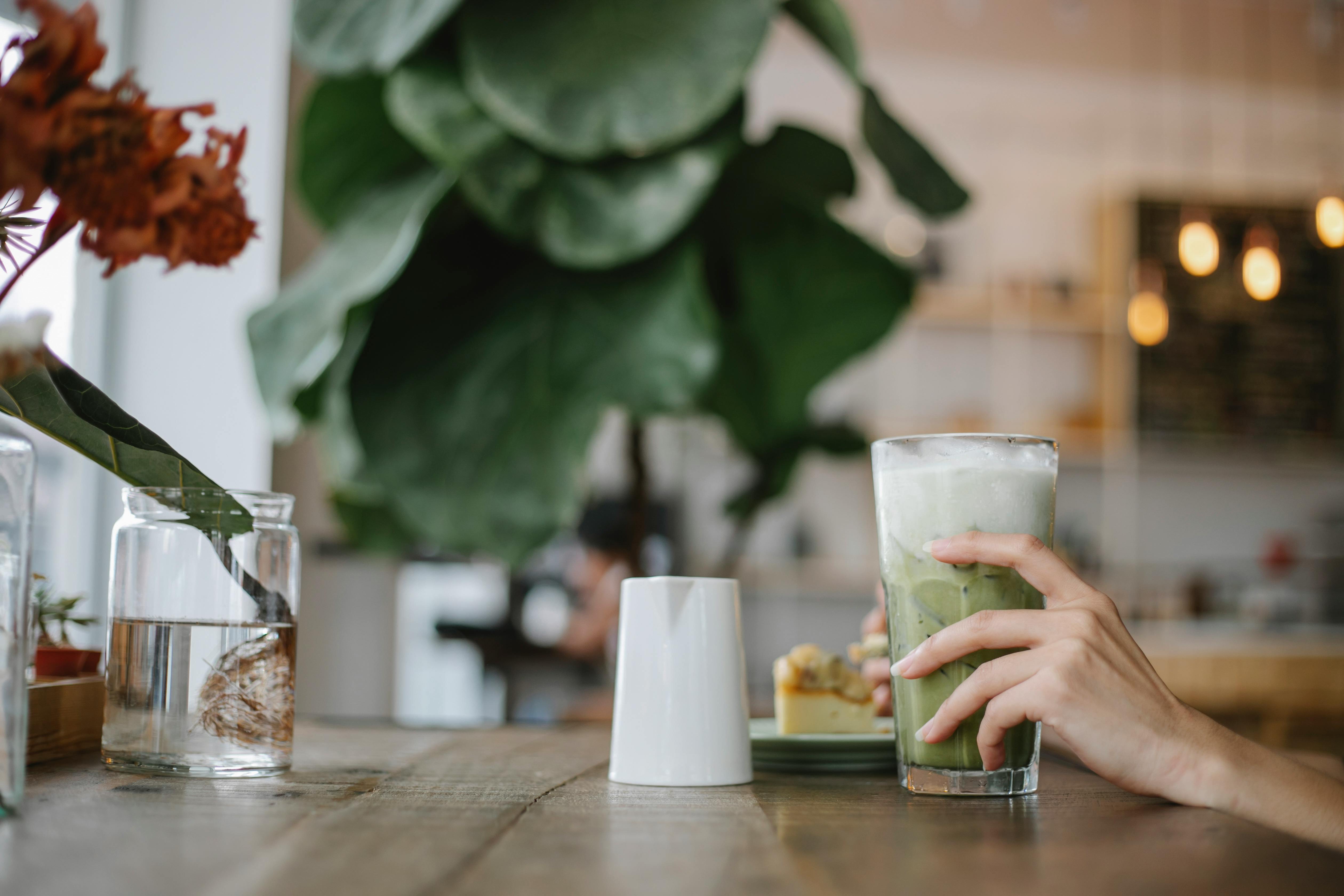 Crop faceless female enjoying iced matcha tea latte and sweet pie while sitting at table in cafeteria