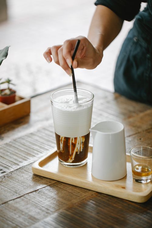 Free Crop anonymous female barista stirring delicious iced coffee latte with straw served on tray with milk jigger and shot of cognac Stock Photo