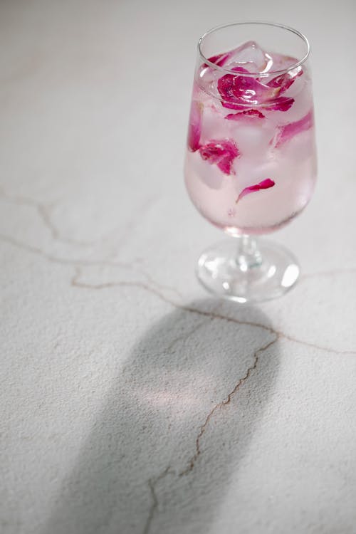 Glass of cold refreshing iced water with pink rose petals served on white table