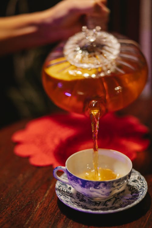 Crop anonymous person pouring aromatic hot tea from crystal glass teapot into ceramic cup on saucer