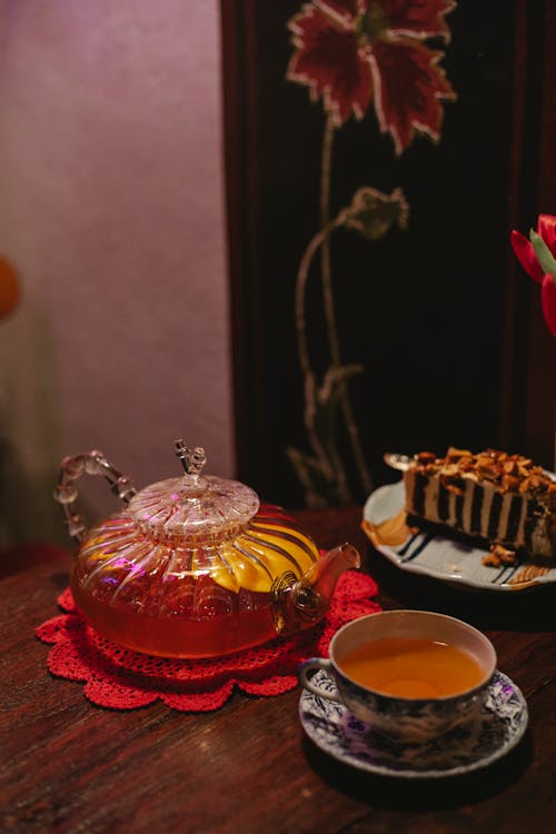 High angle of cup full of hot aromatic tea placed on table near crystal teapot and plate with dessert in cafe